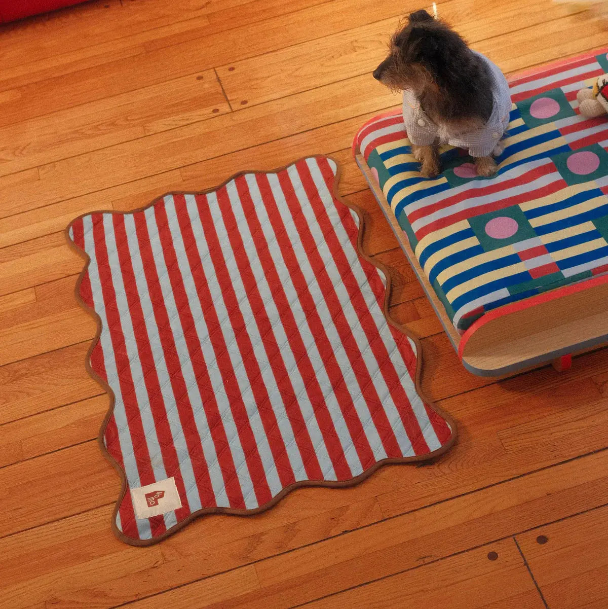 The Ellie Pad washable dog pee pad by NomoHaus placed on wooden floor beside small dog on bed